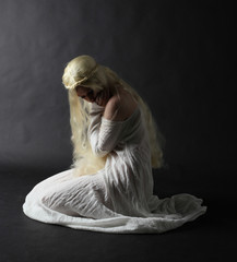 Fototapeta na wymiar Ghostly full length portrait of a woman with long blonde hair wearing a white robe. Standing pose against a black studio background. 