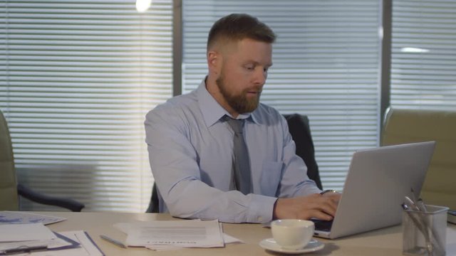 Chest-up shot of red-haired Caucasian corporate lawyer in shirt and tie sitting alone in office late at night, with closed blinds, studying contracts and working on laptop