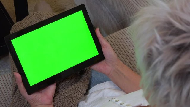 A middle-aged woman sits on a couch in an apartment and looks at a tablet with a green screen - closeup on the tablet