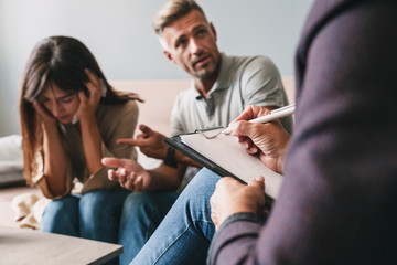 Photo of upset irritated couple having conversation with psychologist on therapy session in room