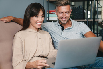 Photo of joyful caucasian couple smiling while watching laptop on sofa at home