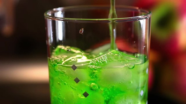 Green estragon lemonade pouring into cocktail glass with ice cubes. Put down blue cocktail straw into drink. Full HD 60fps macro footage with shallow depth and blurring bokeh.