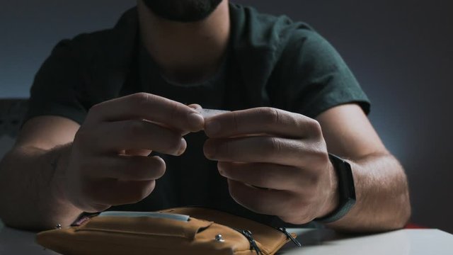 Pan shot of young man's hands close up while rolling up a cigarette, tobacco holder and tabacco accessories on the table foreground
