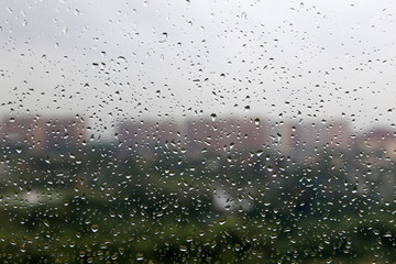 Water drops on the window glass on blurred background of city buildings and cloudy sky. Beautiful raindrops, rainy weather