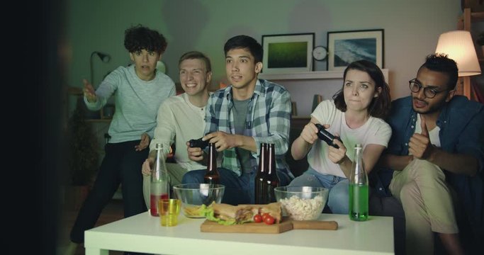 Multiracial group of students are having fun with video game in dark apartment talking laughing sitting on couch holding joysticks. Friendship and leisure concept.