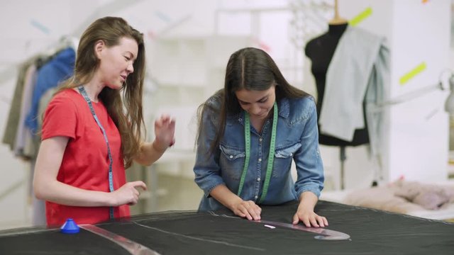 Tracking shot of professional tailor teaching young woman how to use sewing curve rulers to draw pattern on fabric with chalk during individual class