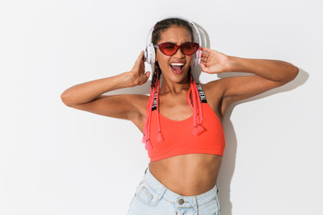 Positive optimistic young african stylish woman in sunglasses posing isolated over white wall background listening music with headphones.