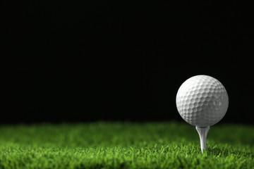 Golf ball with tee on artificial grass against black background, space for text