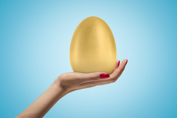 Female hand holding big golden egg on blue background