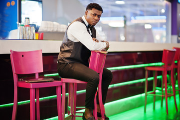 Young handsome african man wearing white shirt, black vest and bow tie posed against bar counter at night club.