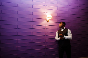 Young handsome african man wearing white shirt, black vest and bow tie posed against purple violet wall.