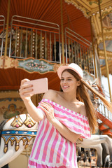 Fototapeta na wymiar Young pretty woman taking selfie near carousel in amusement park
