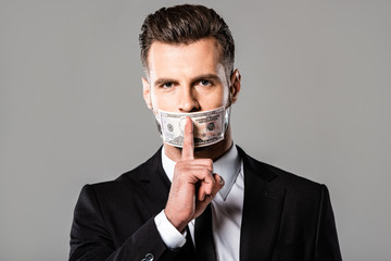 businessman in black suit with dollar banknote on mouth showing hush gesture isolated on grey