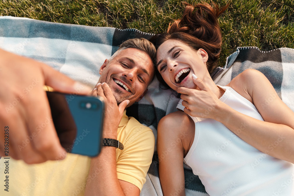 Canvas Prints Cheerful optimistic adult loving couple lies outdoors in a beautiful green nature park take a selfie by mobile phone.