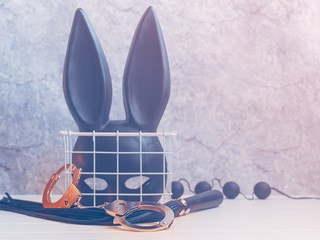 A black mask with rabbit ears is in a white metal basket. Nearby on a white wooden surface are sex...