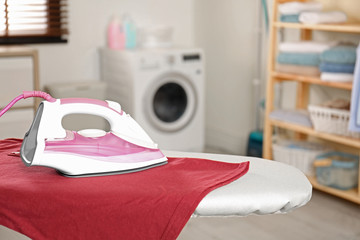 Modern electric iron and clean t-shirt on board in laundry room. Space for text