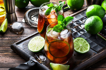 Cuba libre alcohol cocktail with golden rum, lemon juice, cola, lime and ice, dark bar counter background, bar tools, place for text