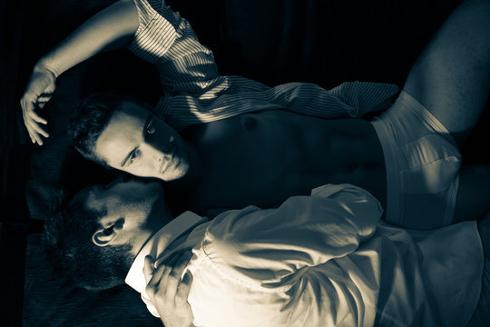 Gay male couple lying in bed with one of them looking at camera as other is holding him