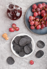 Black potato chips with charcoal, balsamic vinegar in glass, red grapes on a blue ceramic plate on a gray concrete background. Top view.