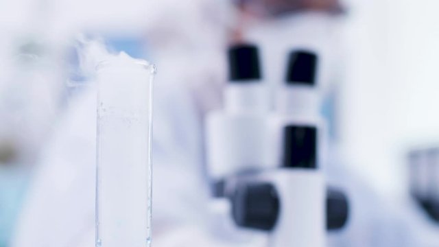 Close up of blue fluid in a bottle smoking in science laboratory. Female scientis in cover all equipemnt in the background.