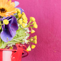 On the one half of the image there is a colorful bunch of flowers in a red pot visible, on the other half of the image, in the background, there is a pink wall for copy space. 