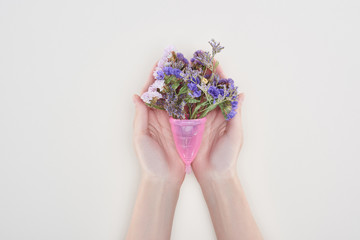 cropped view of woman holding menstrual cup with wildflowers isolated on grey