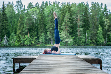Young yogi  girl  practicing yoga, standing in Salamba Sarvangasana exercise, supported Shoulder stand pose on the lake.  Concept of healthy life and natural balance