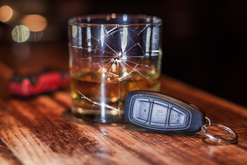A glass of whiskey standing on a table is broken. Nearby are the car keys. In the background is the silhouette of an inverted car. Conceptual photo.