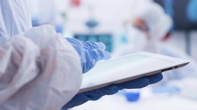 Close up handheld footage of chemist typing on tablet in a research laboratory. Team of scientist working in the background. Scientist in coverall equipment.