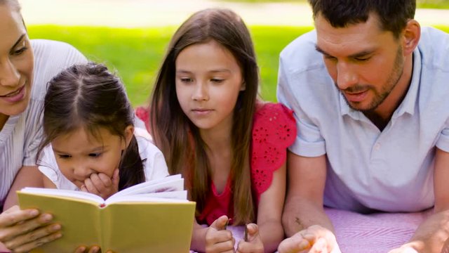 family, leisure and people concept - happy mother, father and two daughters lying on picnic blanket and reading book in summer park