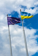 Two flags of Ukraine and the European Union on flagpoles at sky.