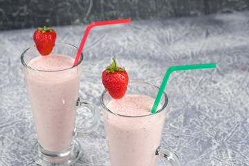 Two glasses with strawberry milkshake with pieces of berries on the edge of the glass on gray background.