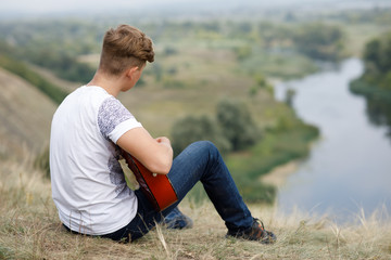 Naklejka na ściany i meble Young boy sits on the hill above the river and plays the guitar