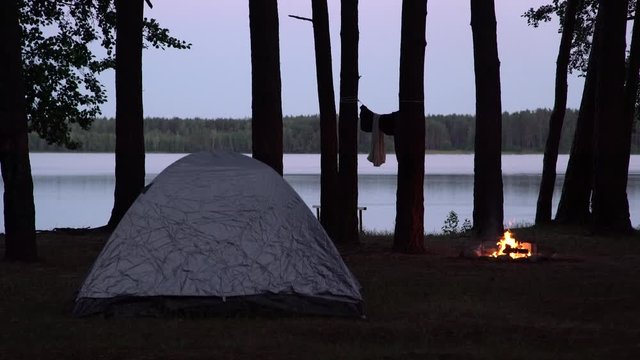 View of the campsite by the lake at twilight. Tent and burning fire place by the water surrounded by trees in the forest. Spending the night in wilderness