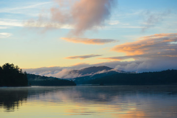 Sun rising over Saranac Lake with the mountains and rolling fog reflecting in the lake; two loons float on the water