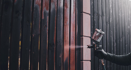 Process of old fence renovation by worker in protective gloves. He is usin airbrush, applying paint.