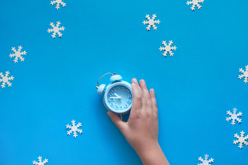 Child's hand holding blue alarm clock showing five to twelve with paper snowflakes