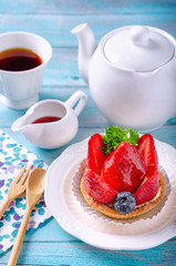 Fruit tart decorated with strawberry and blueberry on a white plate, With a tea of ready-to-drink and a teapot placed on a wooden floor. To feel the atmosphere in the morning.