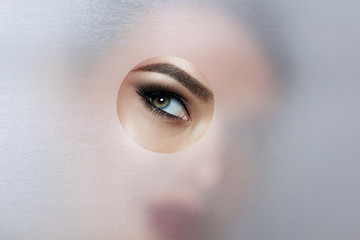 A girl with beautiful green bright eyes with brown shadows and expressive eyebrows looks into the hole of colored paper.