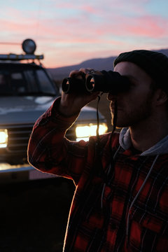 A man with binoculars near the car at sunset. Man Binoculars Looking Mountain Cloudscape Traveling Concept