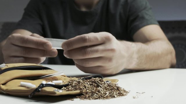 Pan shot of man's hands close up while rolling up a cigarette without filter, tobacco holder and tabacco accessories on the table foreground
