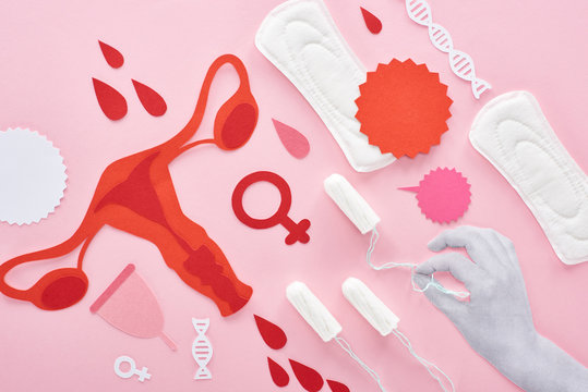 cropped view of white hand holding tampon on pink background with sanitary napkins, paper cut female reproductive internal organs and blood drops