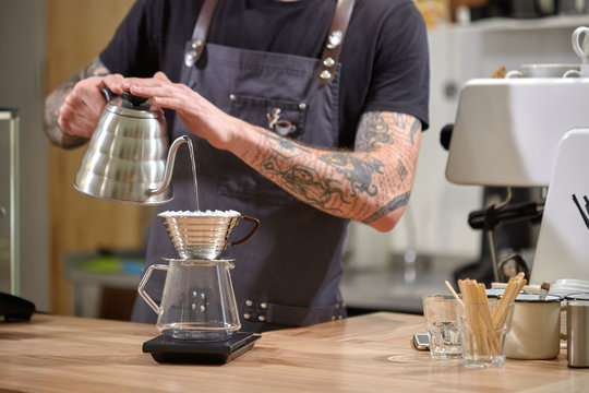 Barista is pouring hot water into the filter cup from coffee pot on a wooden kitchen table.