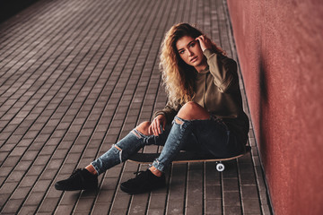 Beautiful young woman in ripped jeans is sitting on her own skateboard at the street.