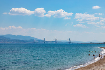 RION / GREECE - JUNE 27, 2019: People swimming on the beach at Gulf of Corinth  near Nafpaktos town. Rion-Antirion Bridge on the background.