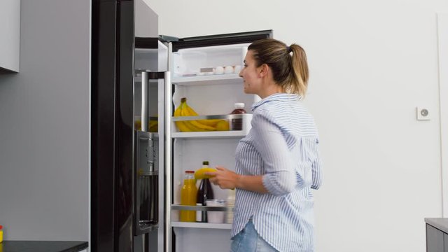 healthy eating, food and diet concept - happy woman opening fridge and taking banana at home kitchen