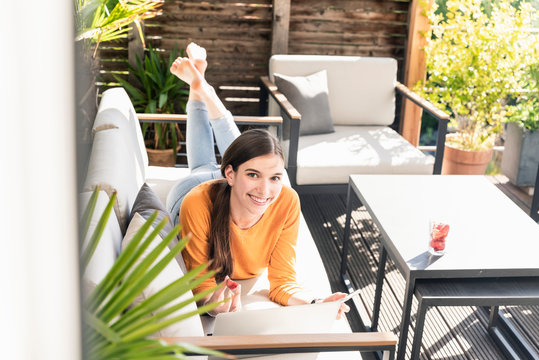 Portrait of smiling young woman lying on couch on terrace using cell phone and laptop - Powered by Adobe