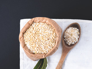 Rice grain in a paper bag and a wooden spoon over black surface. Top view