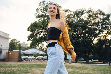 Young pretty cheerful woman in yellow shirt and jeans happily walking on lawn in park
