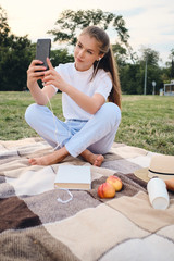 Beautiful teenage girl in earphones dreamily taking photo on cellphone on picnic in park